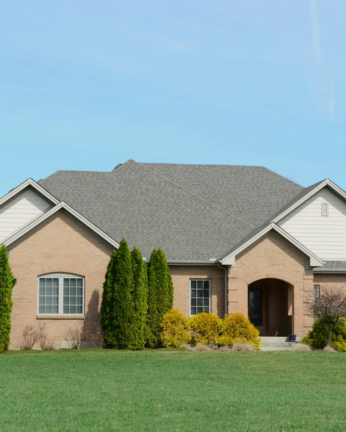 Tan brick single-story suburban home with gray shingle roof, green lawn and blue sky